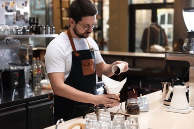 Photo of Barista pouring ground coffee into glass coffeemaker with paper filter at table in cafe