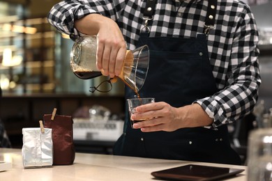 Barista pouring coffee from glass coffeemaker into cup at table in cafe, closeup