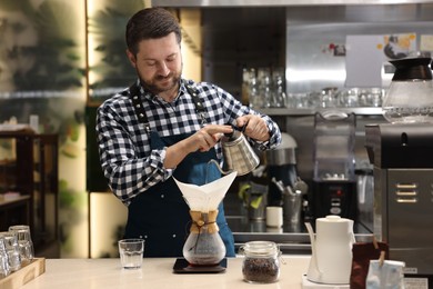 Photo of Barista brewing coffee in glass coffeemaker with paper filter at table in cafe