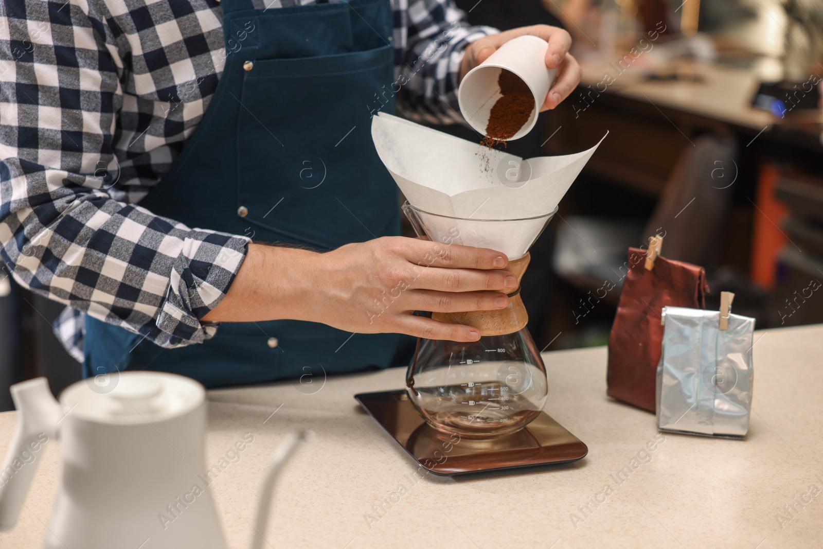 Photo of Barista pouring ground coffee into glass coffeemaker with paper filter at table in cafe, closeup