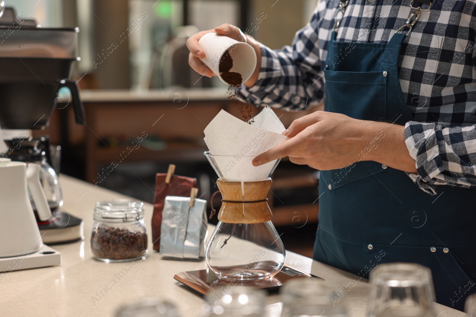 Photo of Barista pouring ground coffee into glass coffeemaker with paper filter at table in cafe, closeup