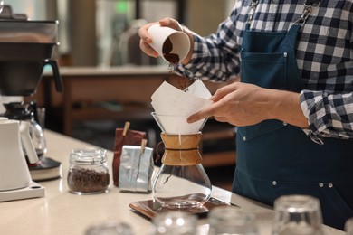 Photo of Barista pouring ground coffee into glass coffeemaker with paper filter at table in cafe, closeup