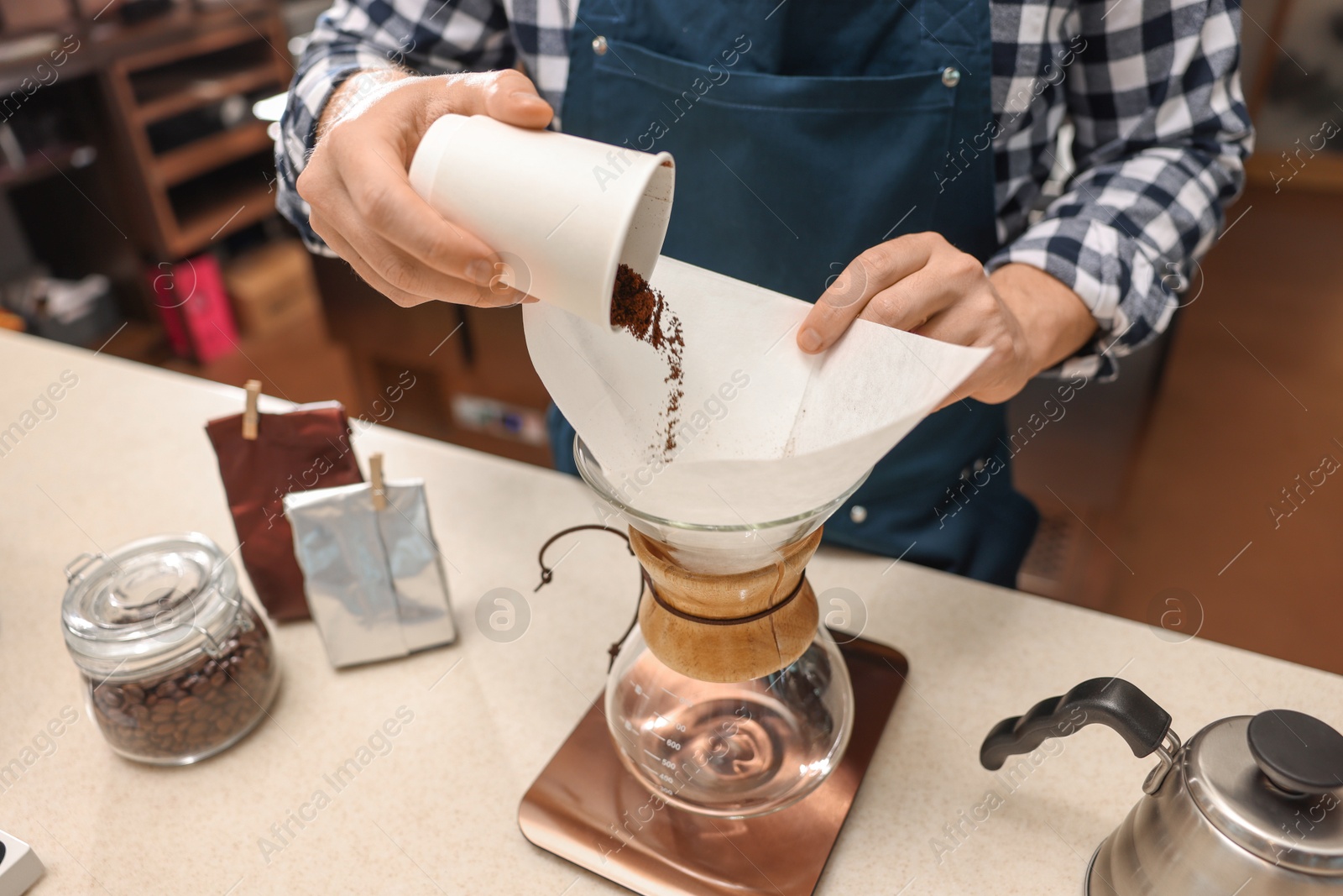 Photo of Barista pouring ground coffee into glass coffeemaker with paper filter at table in cafe, closeup