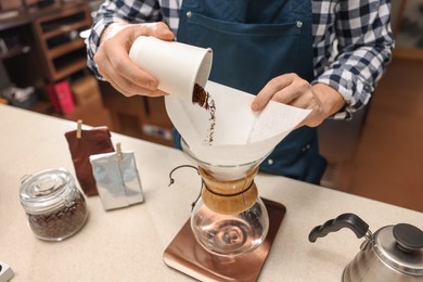 Photo of Barista pouring ground coffee into glass coffeemaker with paper filter at table in cafe, closeup