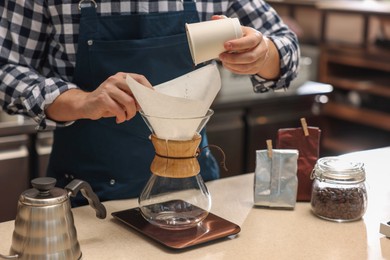 Photo of Barista pouring ground coffee into glass coffeemaker with paper filter at table in cafe, closeup