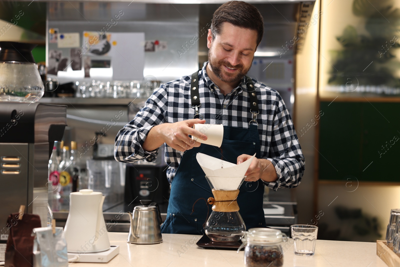 Photo of Barista pouring ground coffee into glass coffeemaker with paper filter at table in cafe