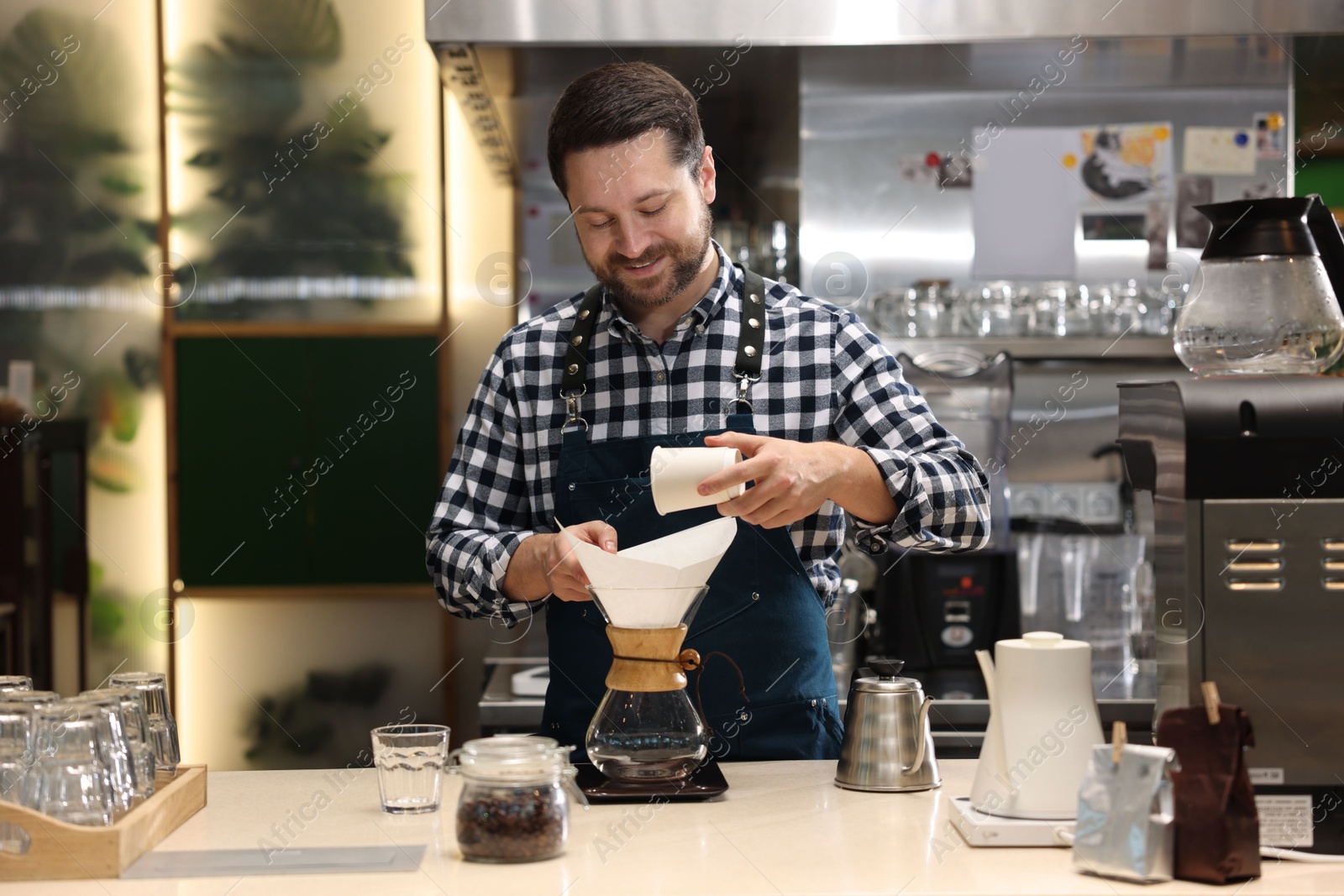 Photo of Barista pouring ground coffee into glass coffeemaker with paper filter at table in cafe