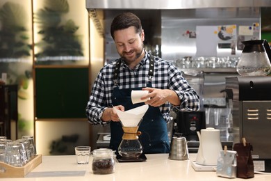 Barista pouring ground coffee into glass coffeemaker with paper filter at table in cafe