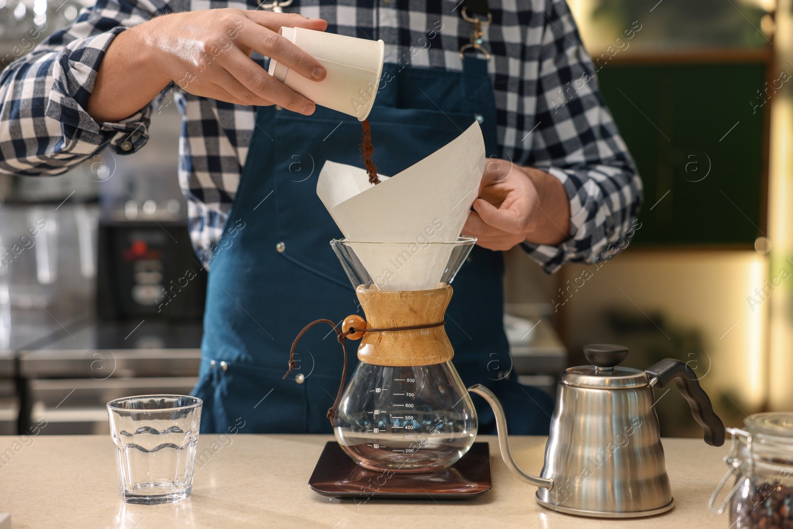 Photo of Barista pouring ground coffee into glass coffeemaker with paper filter at table in cafe, closeup