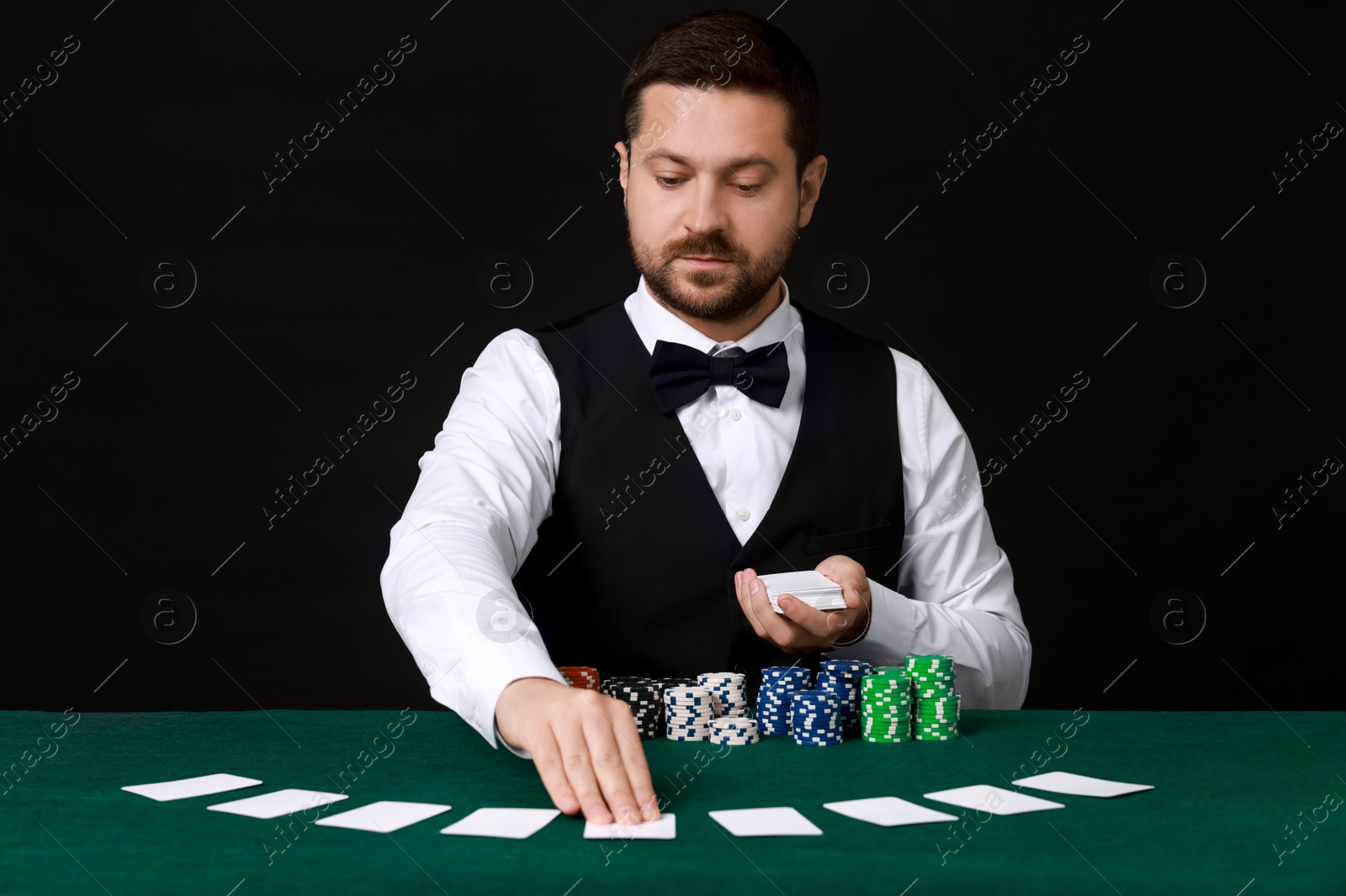 Photo of Professional croupier with playing cards at gambling table against black background