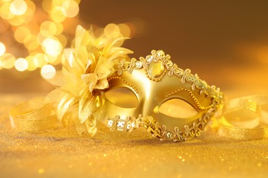 Photo of Beautiful carnival mask on golden shiny surface against blurred lights, closeup