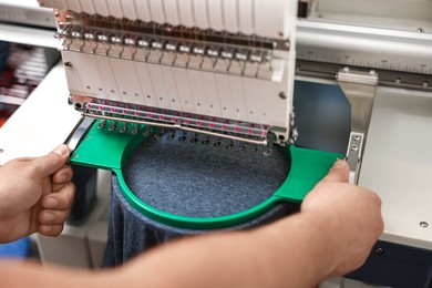 Photo of Man using embroidery machine to make design on T-shirt indoors, closeup