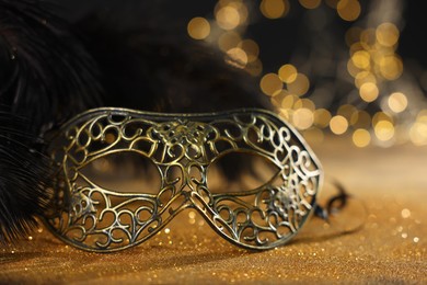 Photo of Beautiful carnival mask and black feathers on golden background against blurred lights, closeup. Bokeh effect