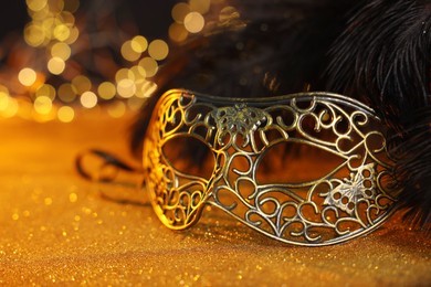 Photo of Beautiful carnival mask and black feathers on golden background against blurred lights, closeup. Bokeh effect