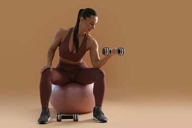 Photo of Woman exercising with dumbbells on fitball against beige background, space for text