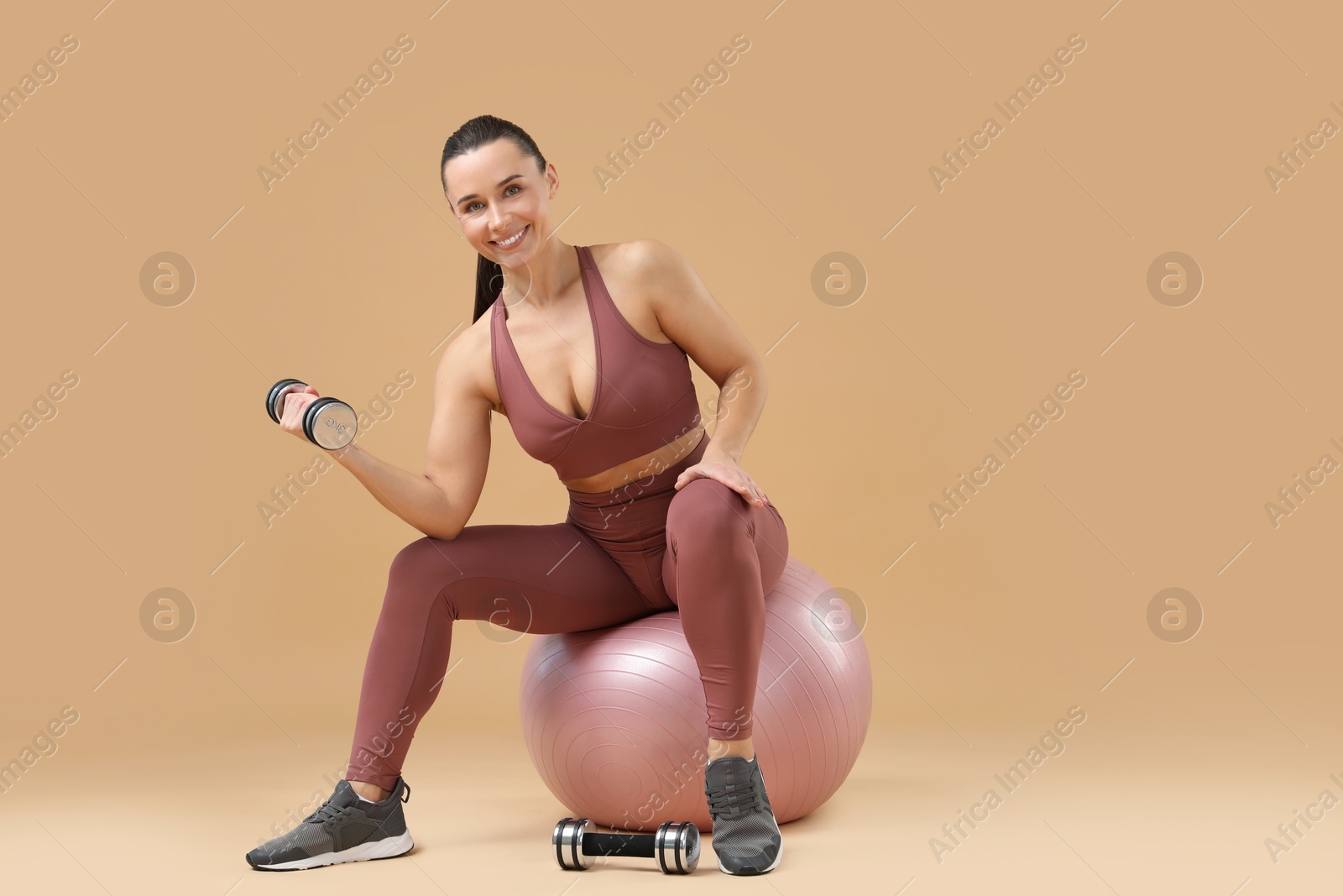 Photo of Woman exercising with dumbbells on fitball against beige background