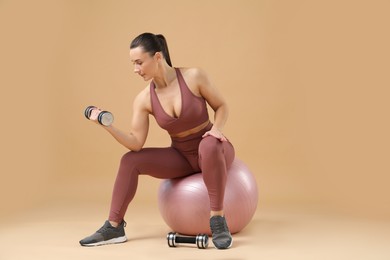 Photo of Woman exercising with dumbbells on fitball against beige background