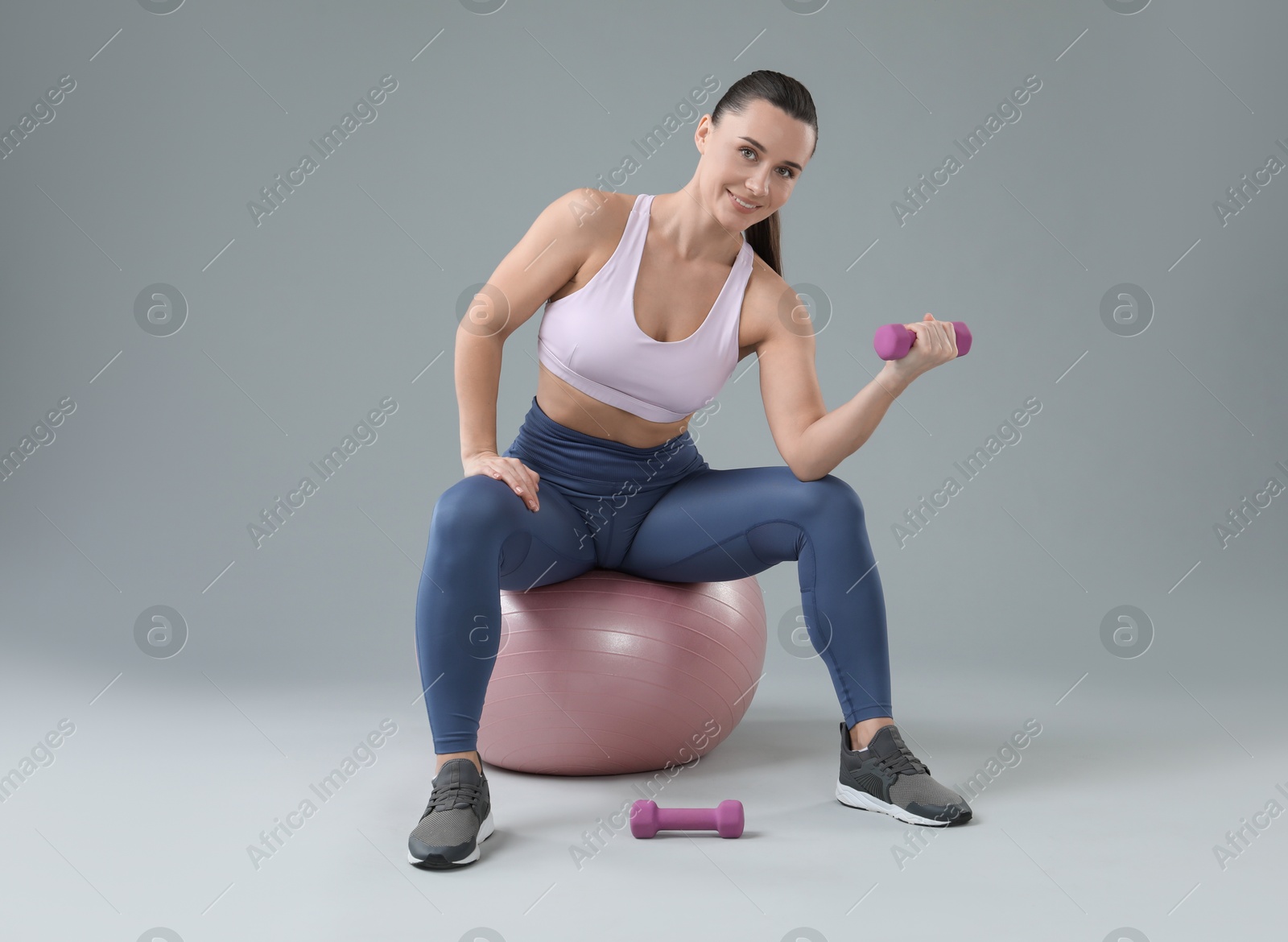 Photo of Woman exercising with dumbbells on fitball against light grey background