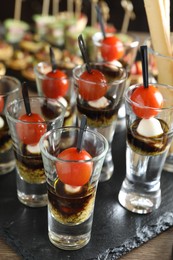 Photo of Tasty canapes with cherry tomatoes, mozzarella, balsamic vinegar and pesto sauce in shot glasses on wooden table, closeup