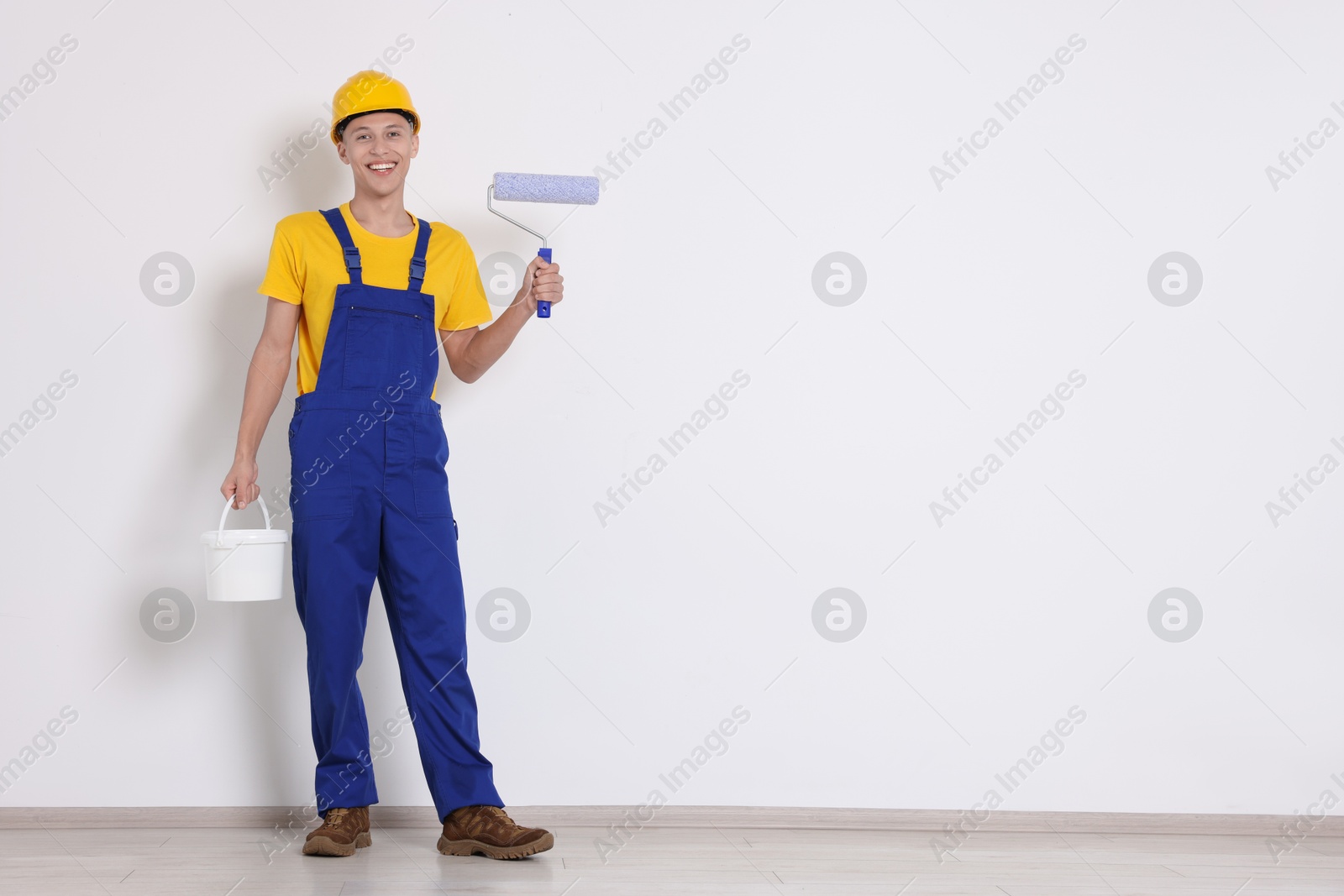 Photo of Young decorator with paint roller and bucket near white wall, space for text