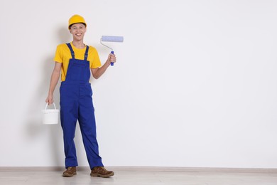 Young decorator with paint roller and bucket near white wall, space for text