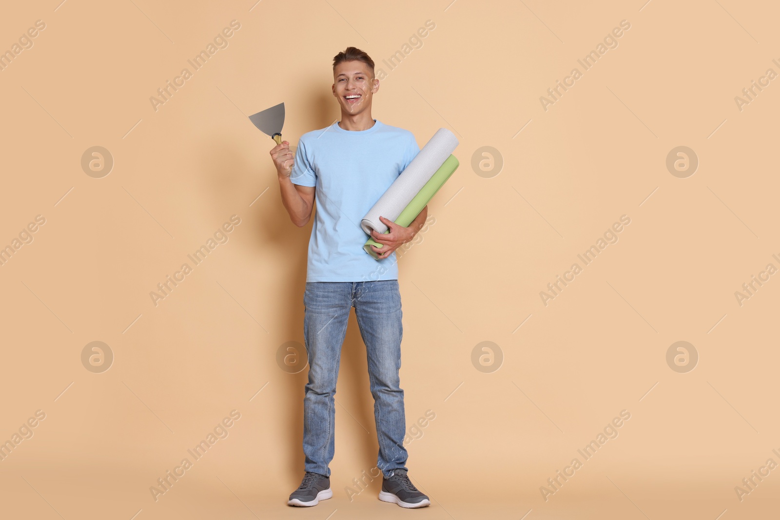 Photo of Young decorator with putty knife and rolls of wallpaper on beige background