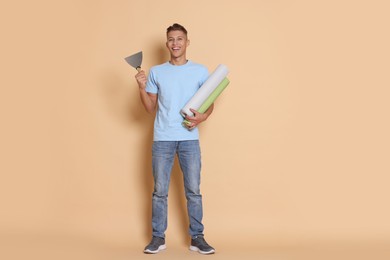 Young decorator with putty knife and rolls of wallpaper on beige background