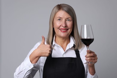 Senior woman with glass of wine showing thumbs up on grey background