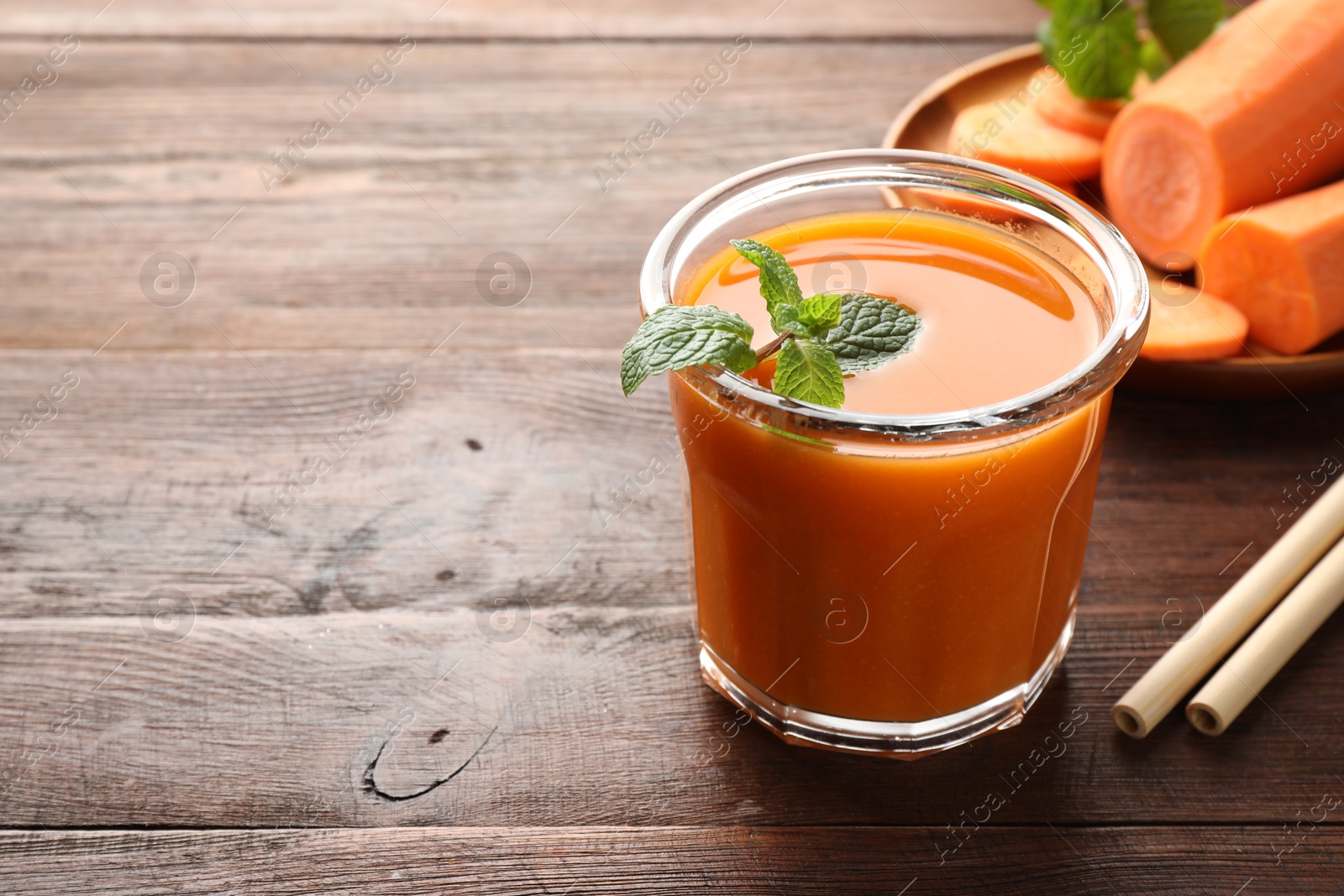 Photo of Fresh carrot juice in glass, vegetables, mint and straws on wooden table, space for text