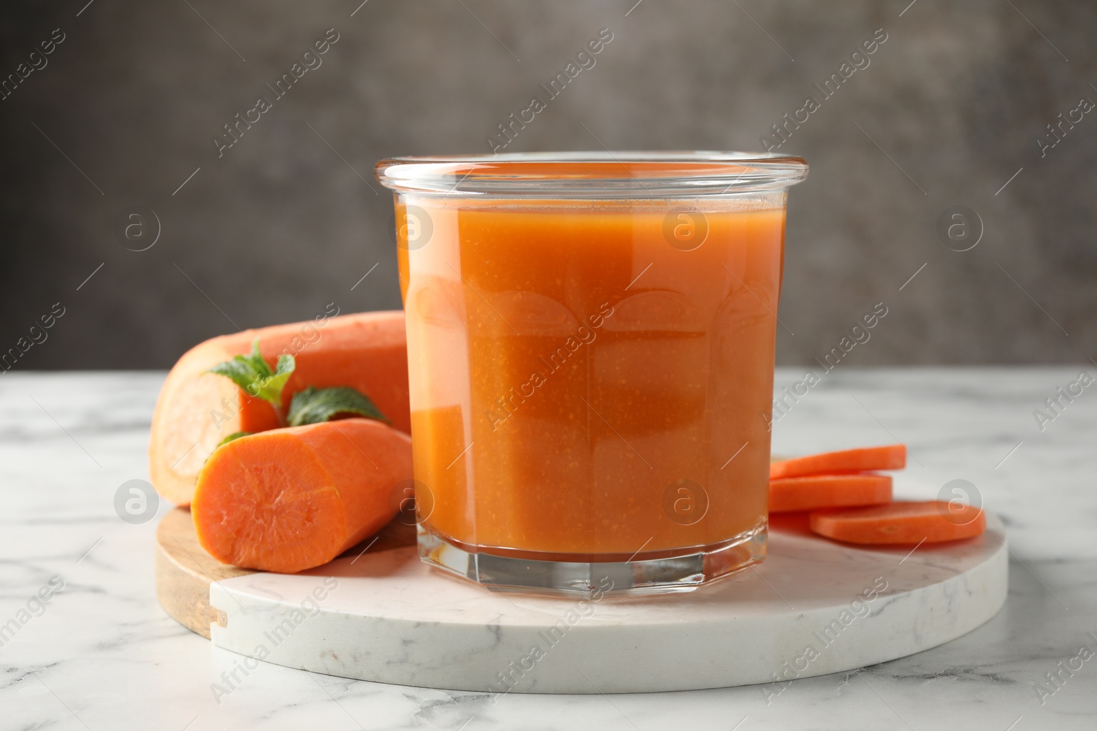 Photo of Fresh carrot juice in glass and vegetables on white marble table against gray background, closeup