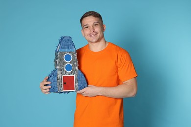 Photo of Happy man with pinata in shape of spaceship on light blue background