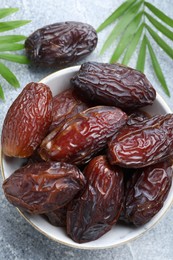 Photo of Many tasty dried dates in bowl and leaves on gray textured table, flat lay