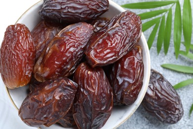 Photo of Many tasty dried dates in bowl and leaves on gray textured table, flat lay