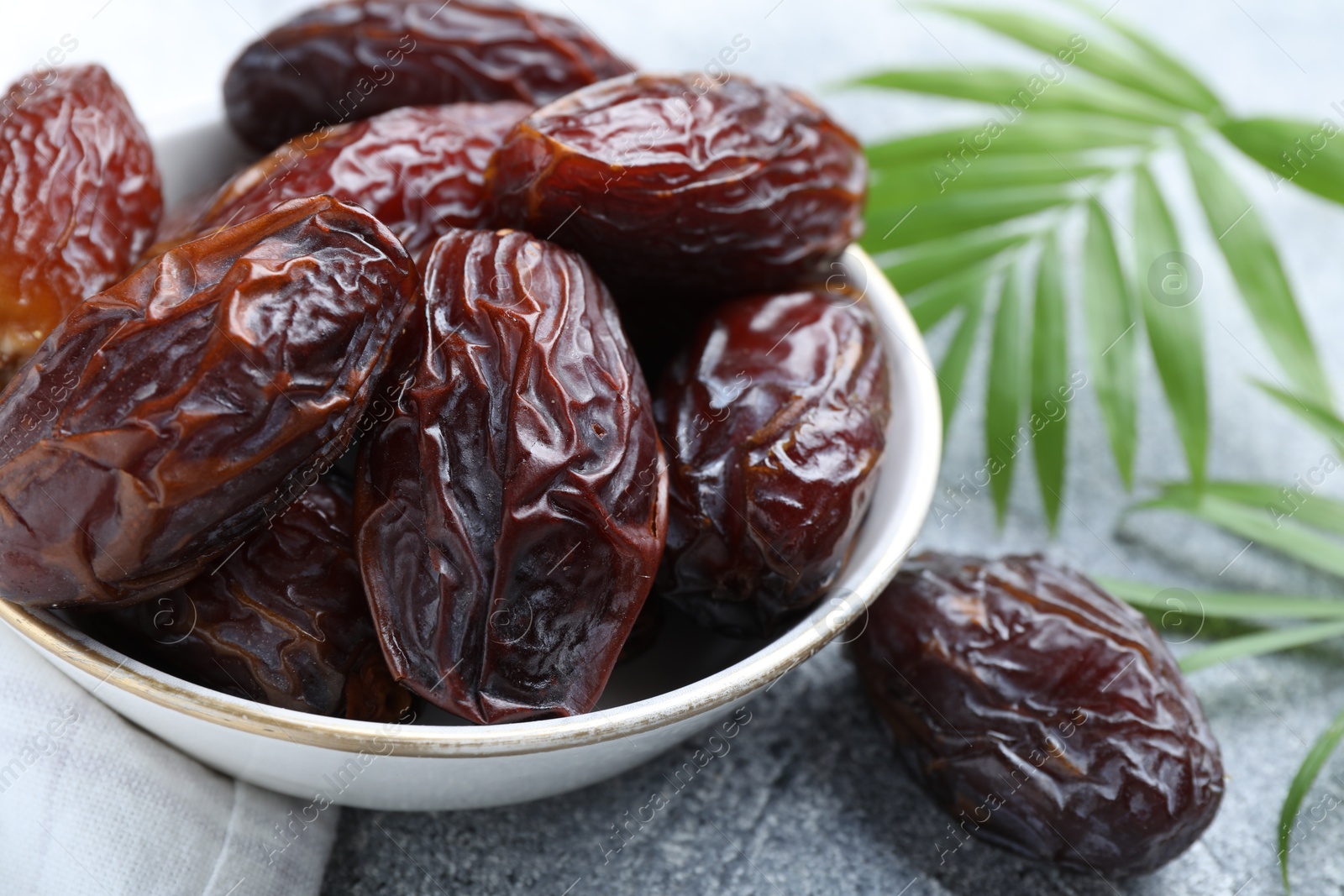 Photo of Many tasty dried dates in bowl and leaves on gray textured table, closeup