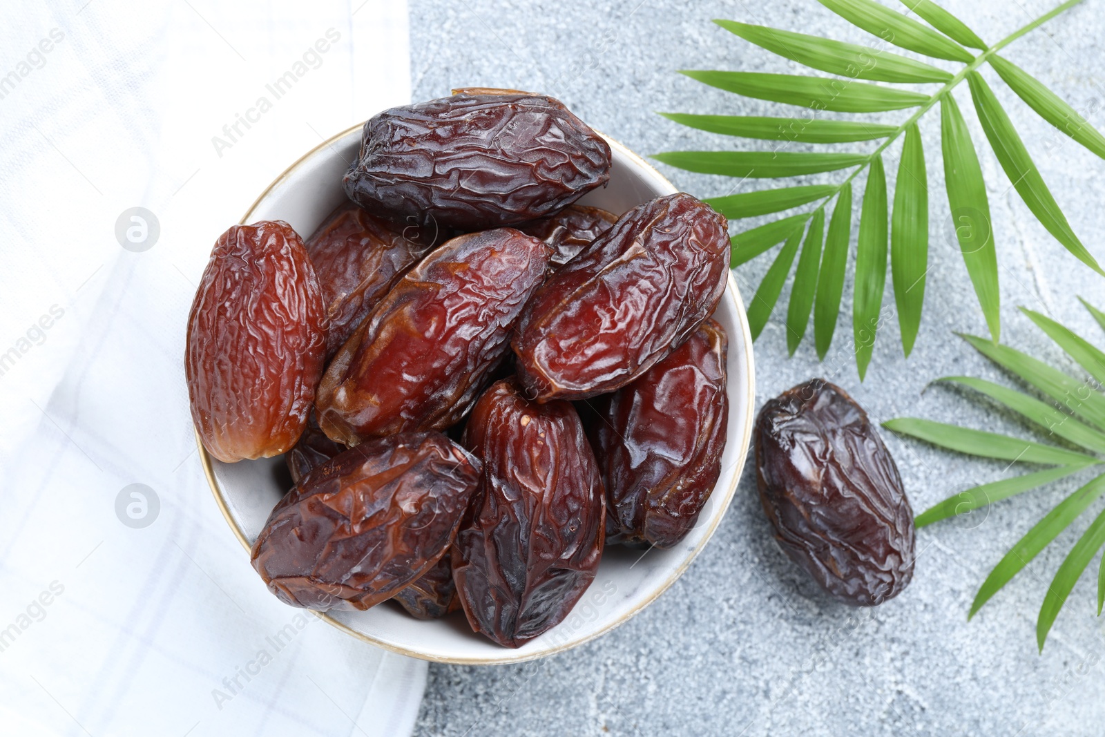 Photo of Many tasty dried dates in bowl and leaves on gray textured table, flat lay