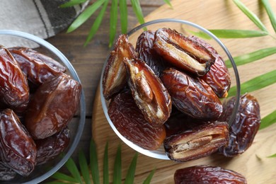 Photo of Many tasty dried dates in bowls and leaves on wooden table, flat lay