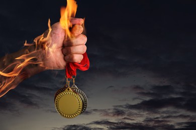 Image of Man holding gold, silver and bronze medals in his flaming hand against darkened sky, closeup