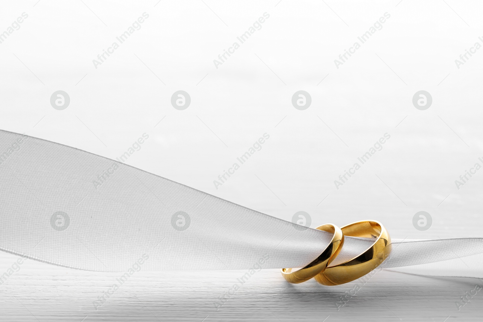 Photo of Beautiful golden wedding rings and white ribbon on light wooden background, space for text