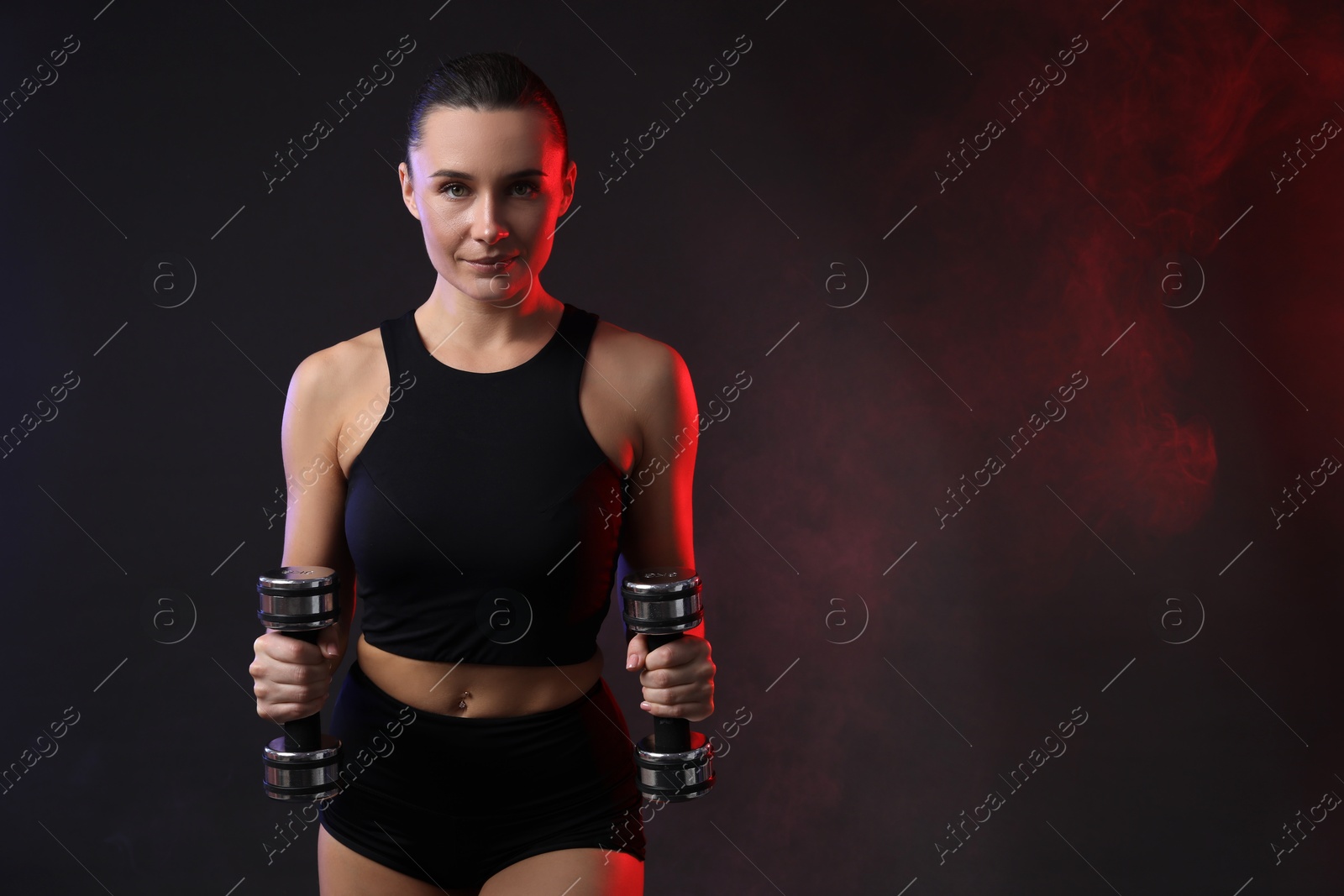 Photo of Woman exercising with dumbbells in smoke on dark background, space for text