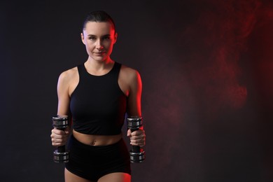 Woman exercising with dumbbells in smoke on dark background, space for text