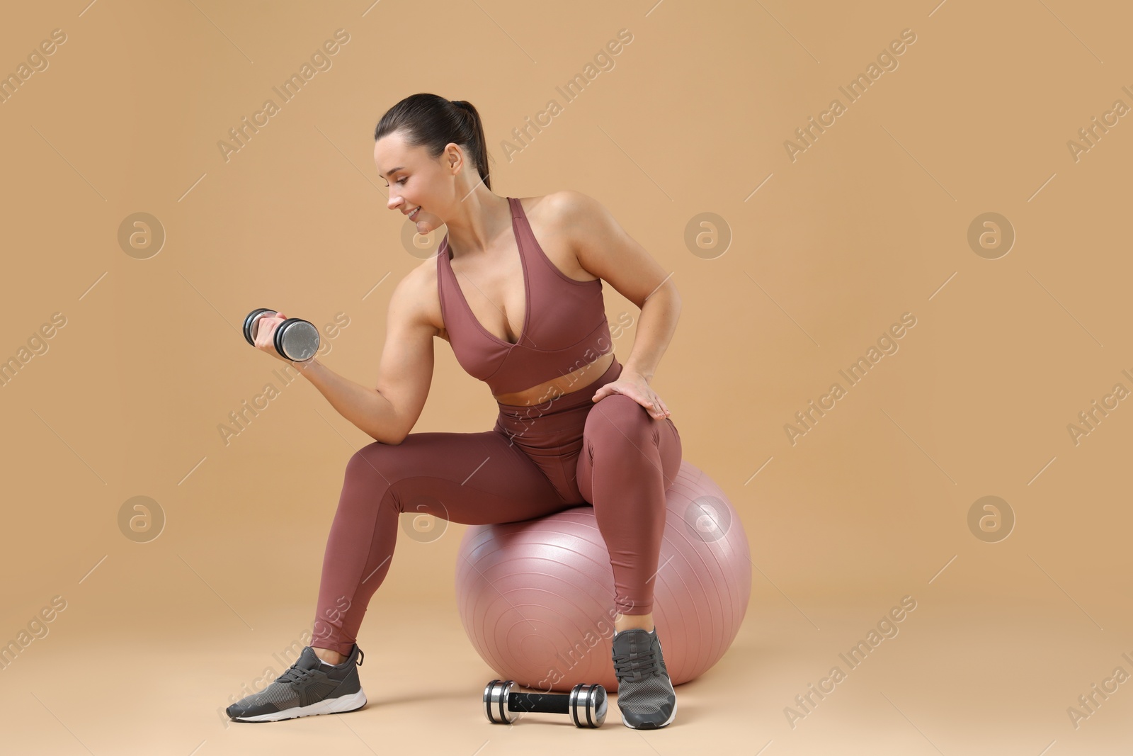 Photo of Woman exercising with dumbbells on fitball against beige background