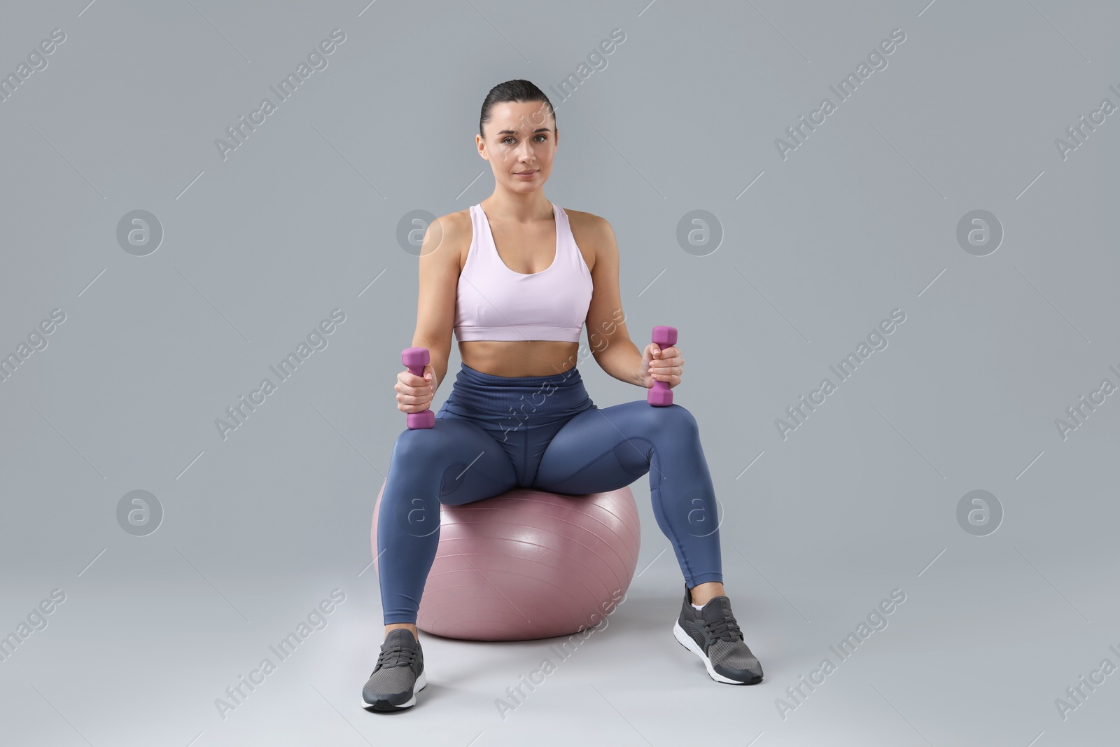 Photo of Woman exercising with dumbbells on fitball against light grey background