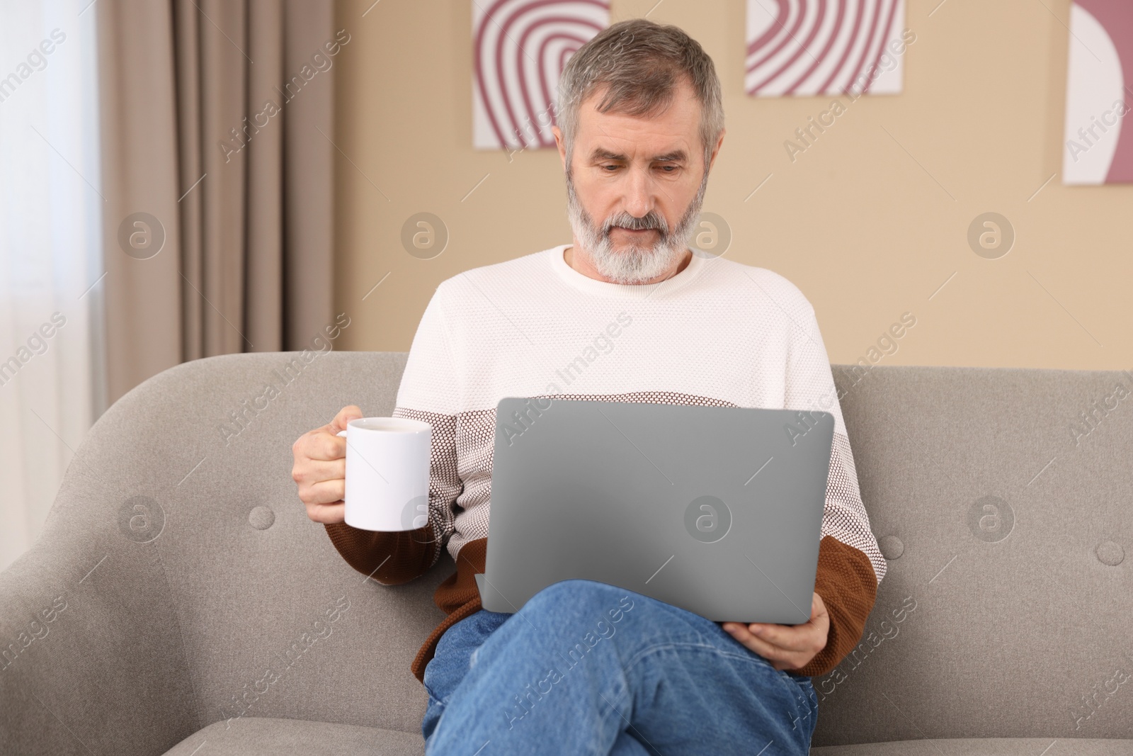 Photo of Mature man with laptop and cup of hot drink on sofa at home