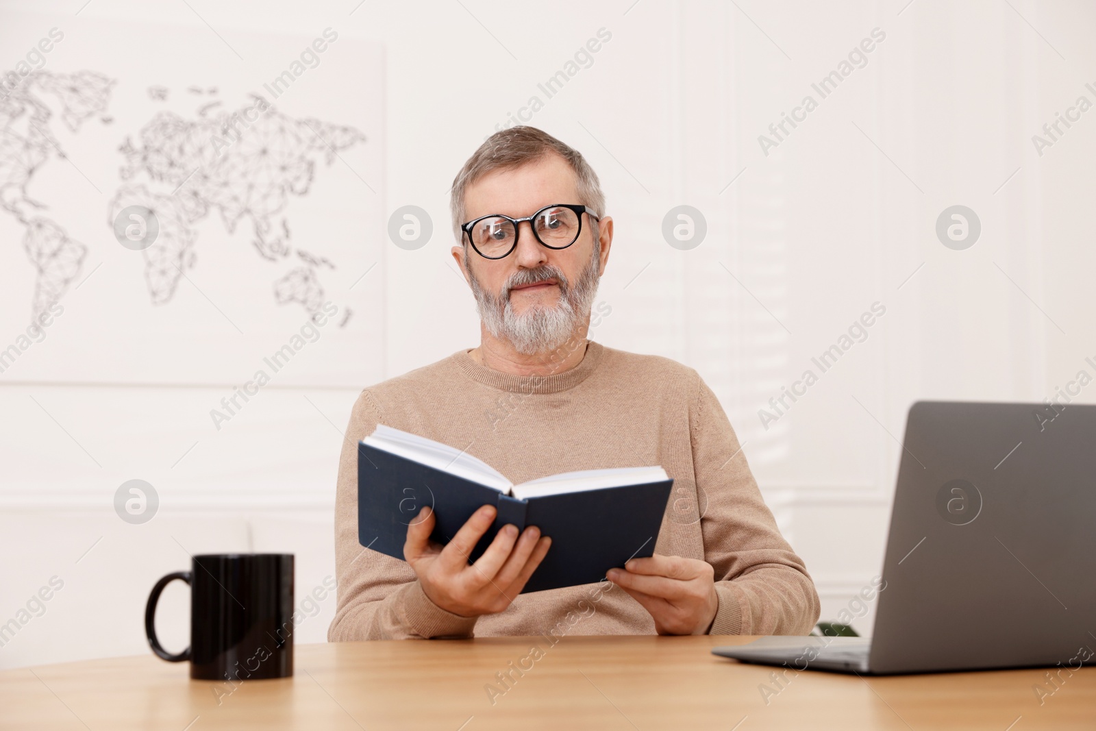 Photo of Mature man reading book at table with laptop indoors