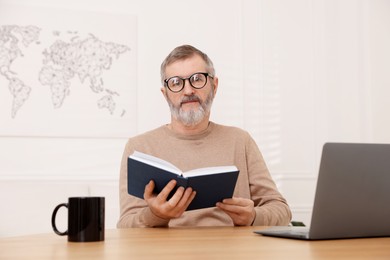 Photo of Mature man reading book at table with laptop indoors