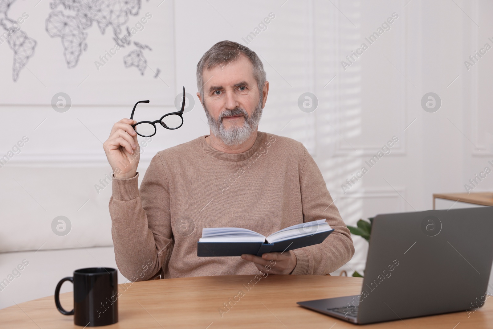 Photo of Mature man reading book at table with laptop indoors