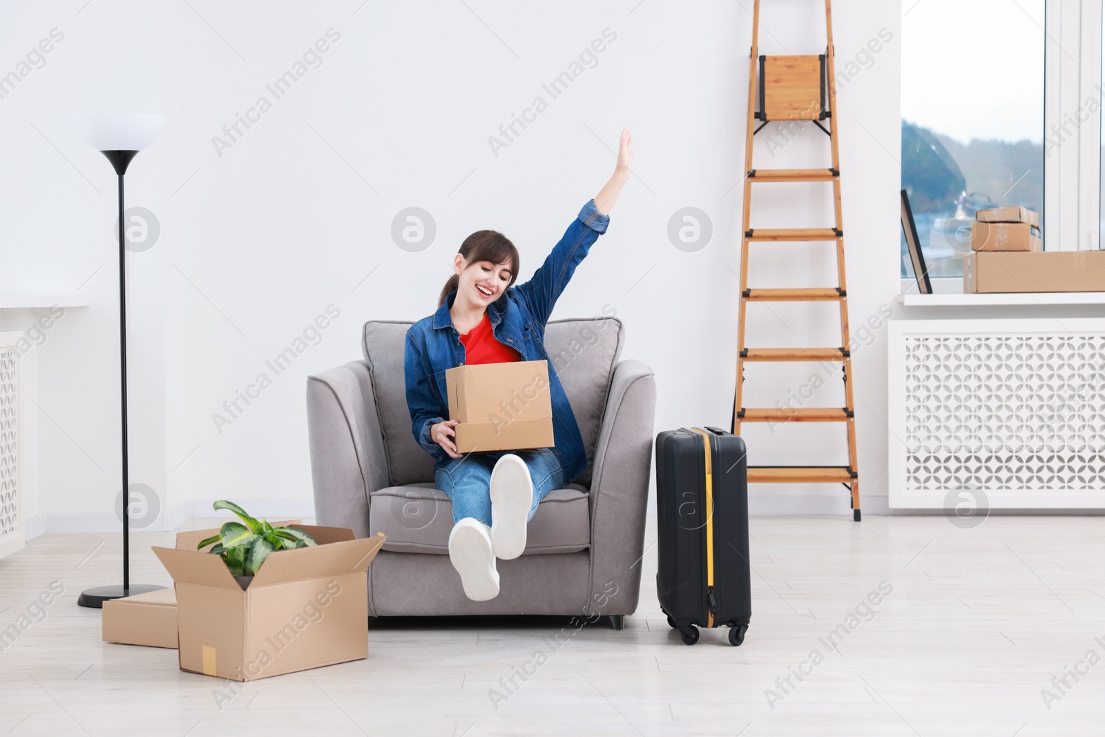 Photo of Happy woman with moving boxes and suitcase in new apartment. Housewarming party