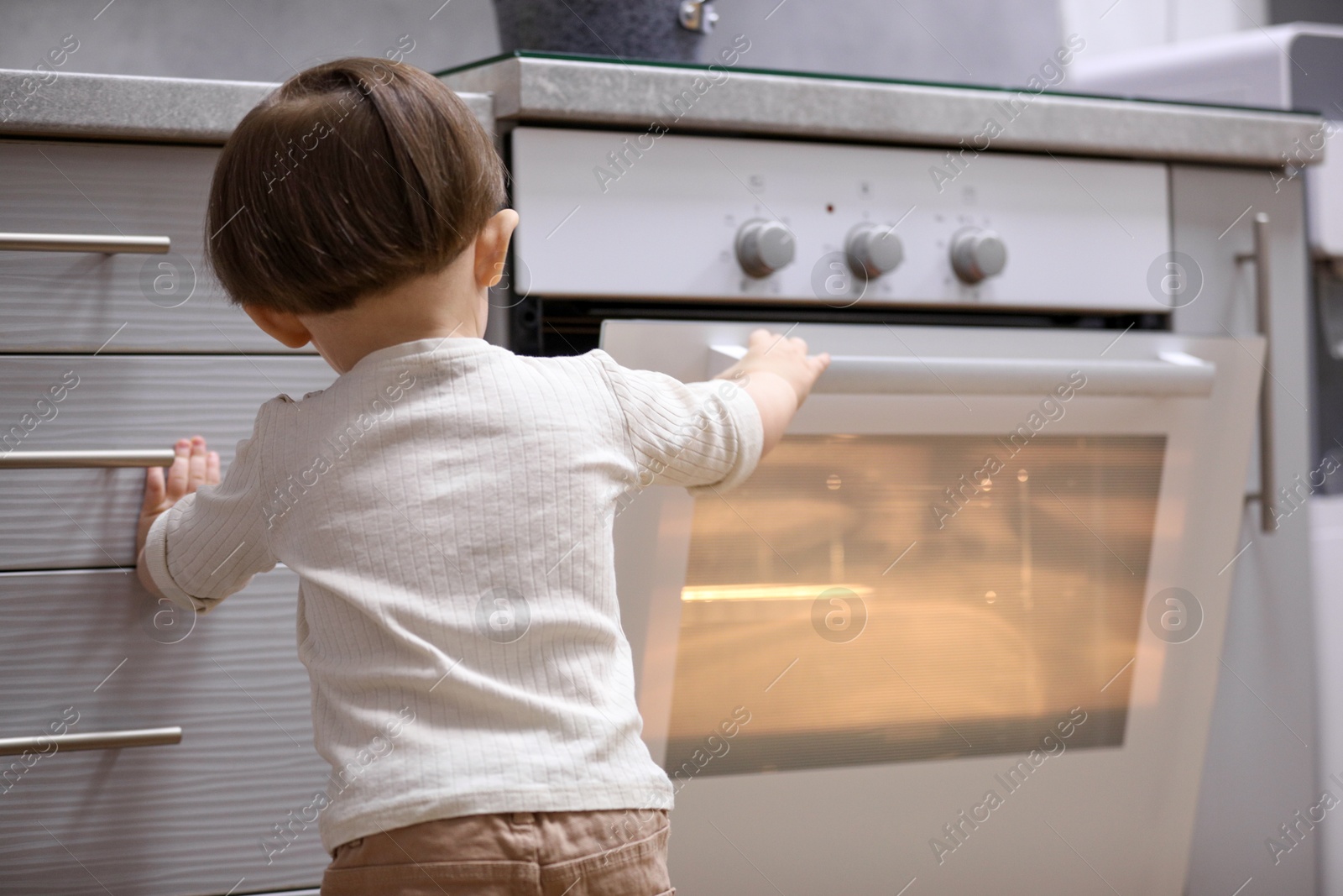 Photo of Little boy playing with oven in kitchen, back view. Dangerous situation