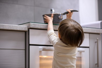 Little boy playing with pot on stove in kitchen, back view. Dangerous situation