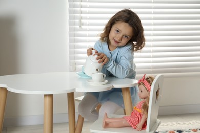 Cute little girl playing tea party with doll at white table in room